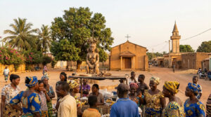 Photo d'un village béninois : statue traditionnelle, église, mosquée et habitants en tenue colorée, illustrant la diversité religieuse.